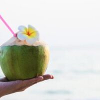 Fresh coconut in hand with plumeria decorated on beach with sea wave background - tourist with fresh fruit and sea sand sun vacation background concept